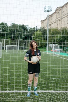 A woman soccer player standing with a ball on a green field, netting in the foreground.