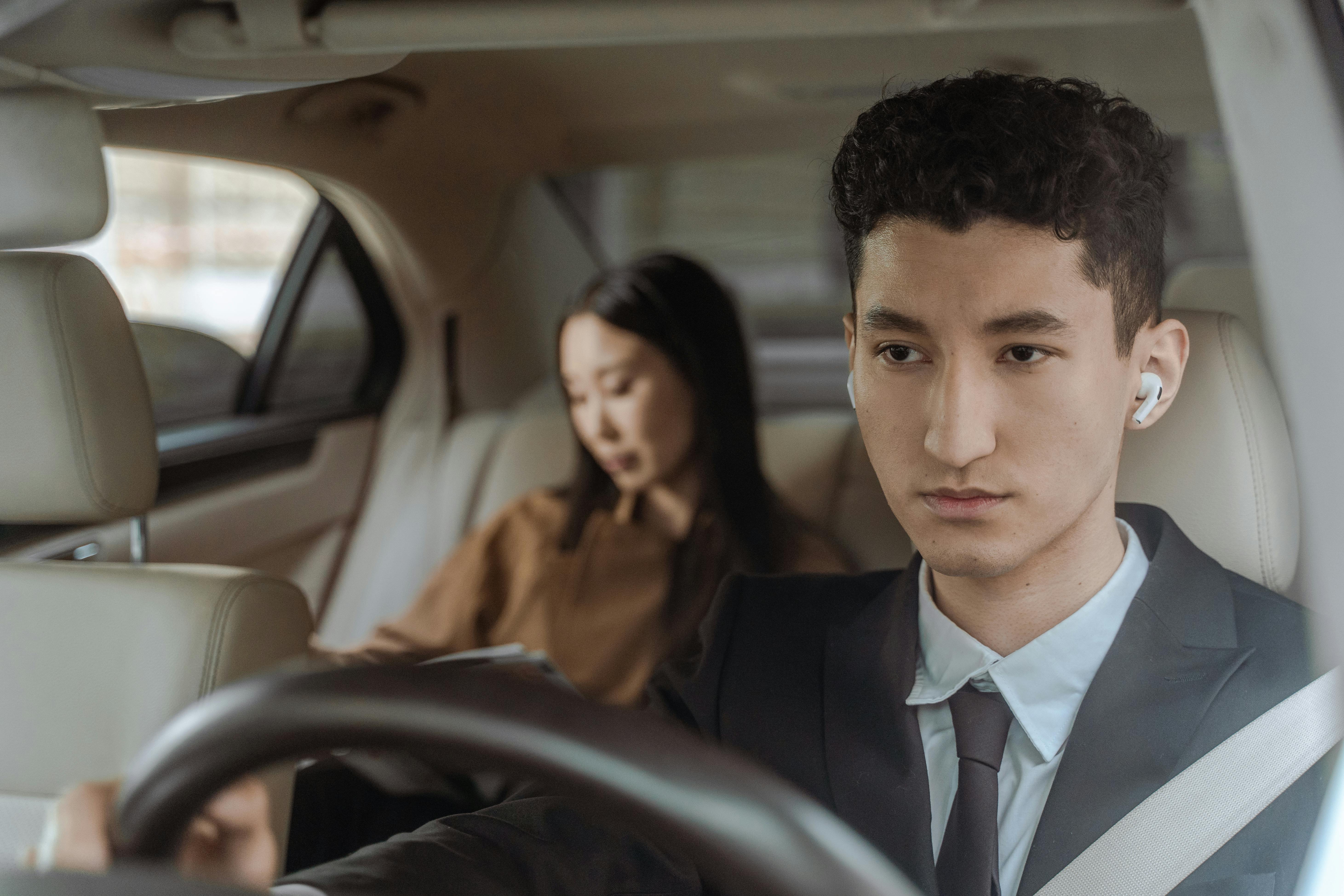 Young professional driver wearing suit and earpiece in luxury car interior with female passenger