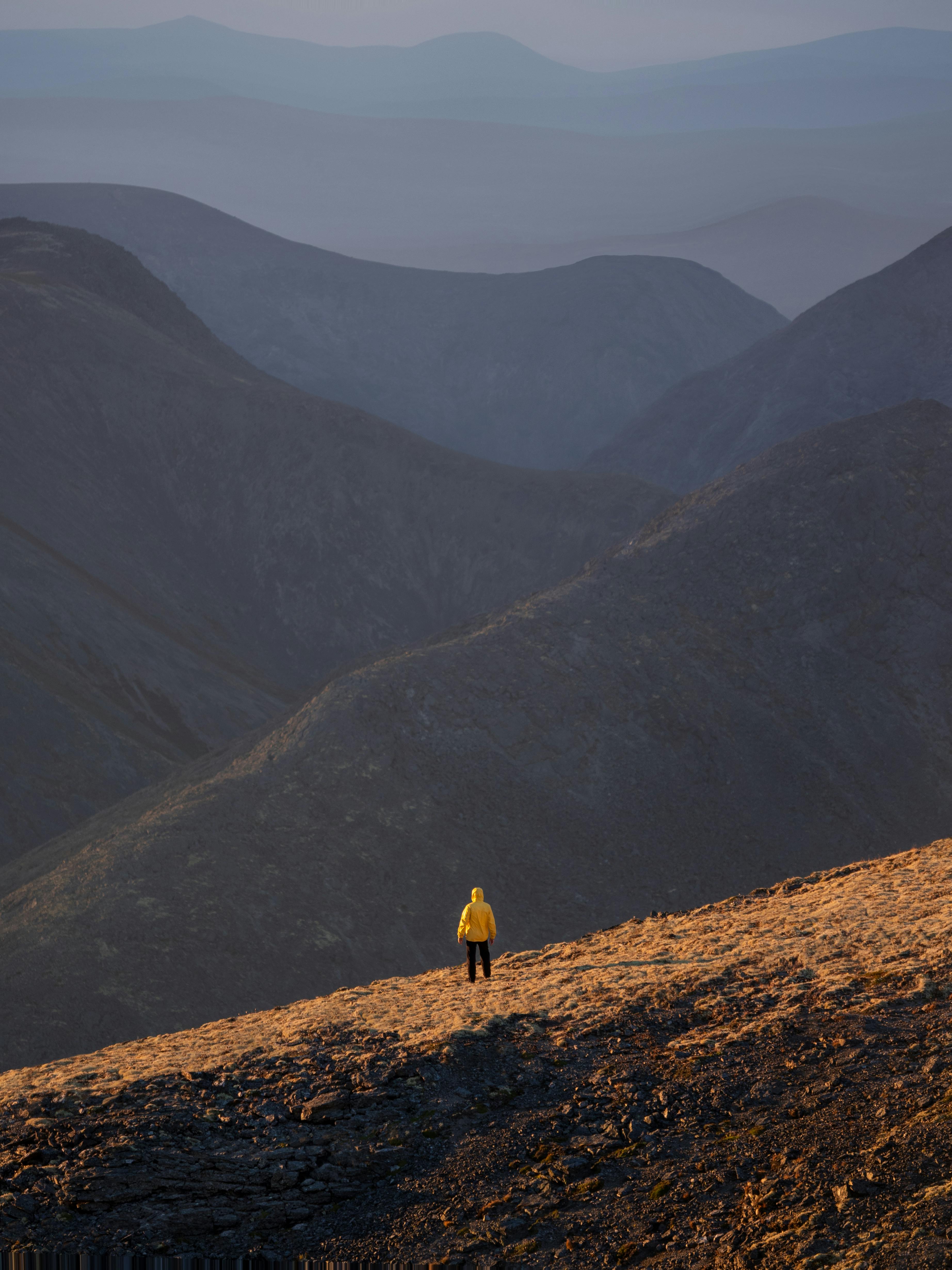 A solitary hiker in a yellow jacket stands amidst the vast Murmansk mountain landscape under a twilight sky.