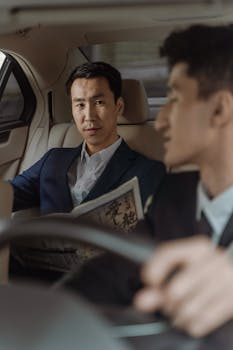 Two men in business suits in a luxury car interior, reading a newspaper during a commute.
