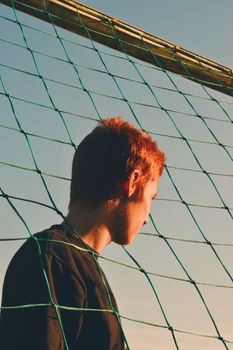 Young adult stands by a soccer net, bathed in warm sunset light, contemplating.