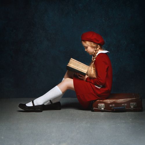 Free A young girl in a red dress and beret sits reading a book, showcasing innocence and style. Stock Photo