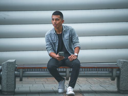 Free Young man in denim jacket sitting on a bench and using smartphone outdoors. Stock Photo