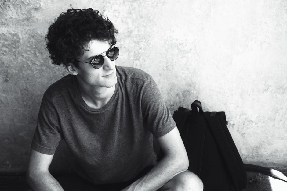 Black and white portrait of a stylish young man wearing sunglasses, sitting casually next to a backpack.