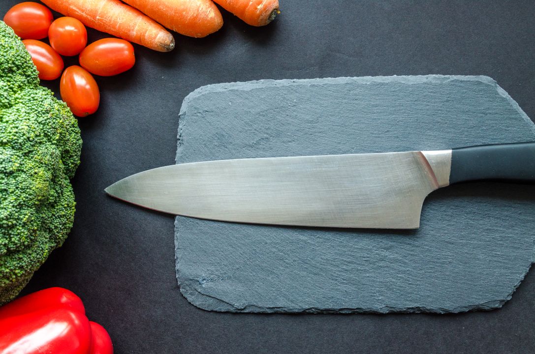 10 Essential Knife Skills Every Home Cook Must Learn 3 A sharp chef's knife rests on a dark slate board surrounded by fresh colourful vegetables — capsicum, carrots, cherry tomatoes — ideal for julienne prep.