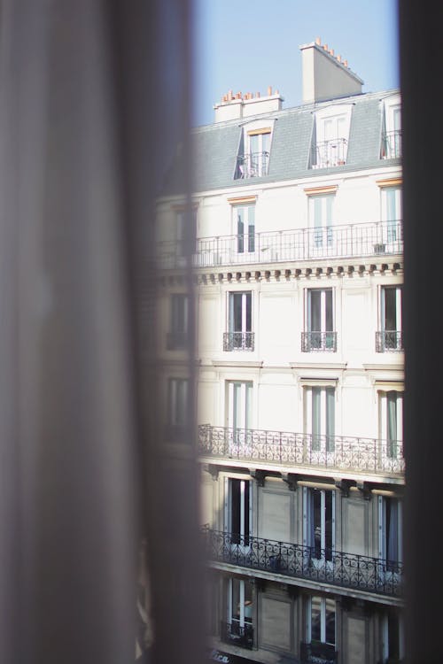 Hotel with elegant balconies and bay windows in Paris as seen from opposite building through slightly ajar window curtains on sunny day