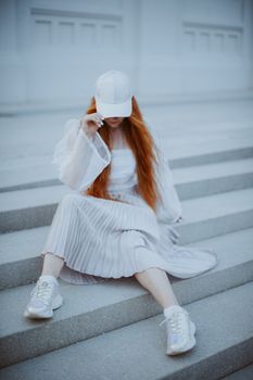 Stylish woman in white outfit and cap sitting gracefully on steps.