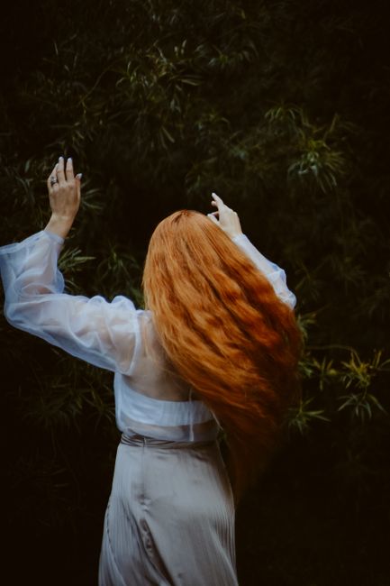 Elegant woman with flowing red hair surrounded by lush greenery, raising her hands in a serene moment.