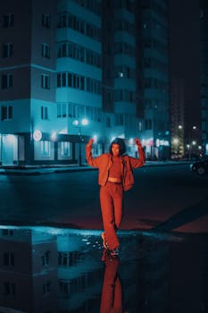 Urban night scene with a woman posing on a city street, illuminated by neon lights reflecting in a puddle.