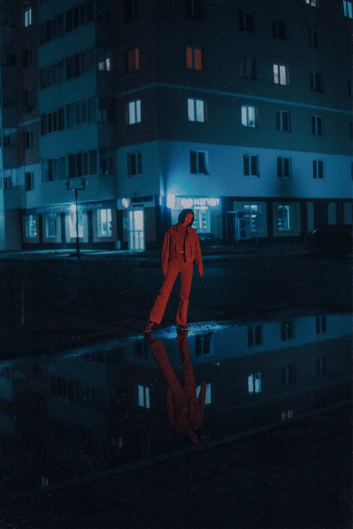 Free A person in red stands in an urban street at night, illuminated by neon lights and reflected in a puddle. Stock Photo