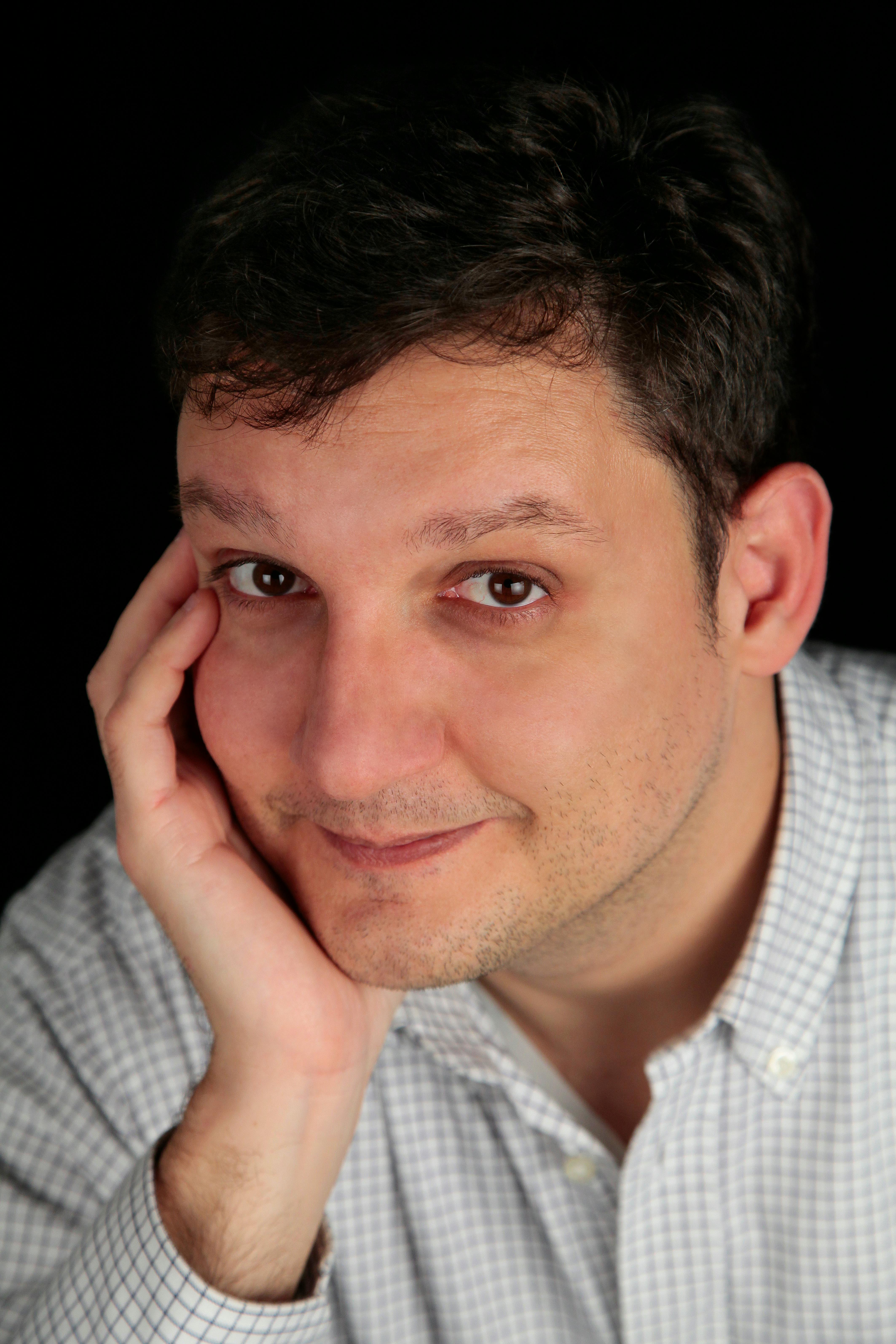 Close-up portrait of a smiling man in a casual shirt, resting his face on his hand against a dark background.