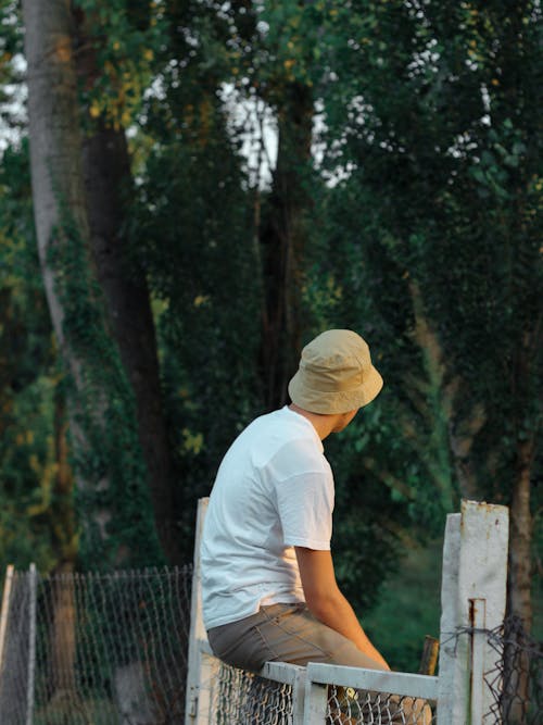 
A Man Wearing a White Shirt and a Bucket Hat Sitting on a Fence