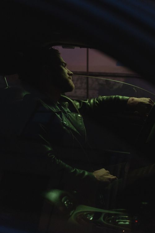 Free A man in a leather jacket sits in a car, driving at night, with a moody atmosphere. Stock Photo