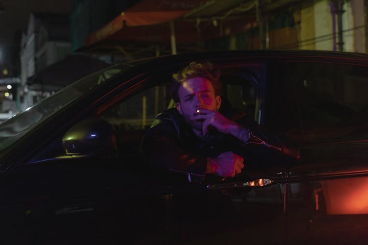 A Man Smoking A Cigarette While Leaning On A Car Window