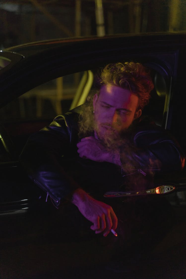 A Man Leaning On A Car Window And Smoking A Cigarette