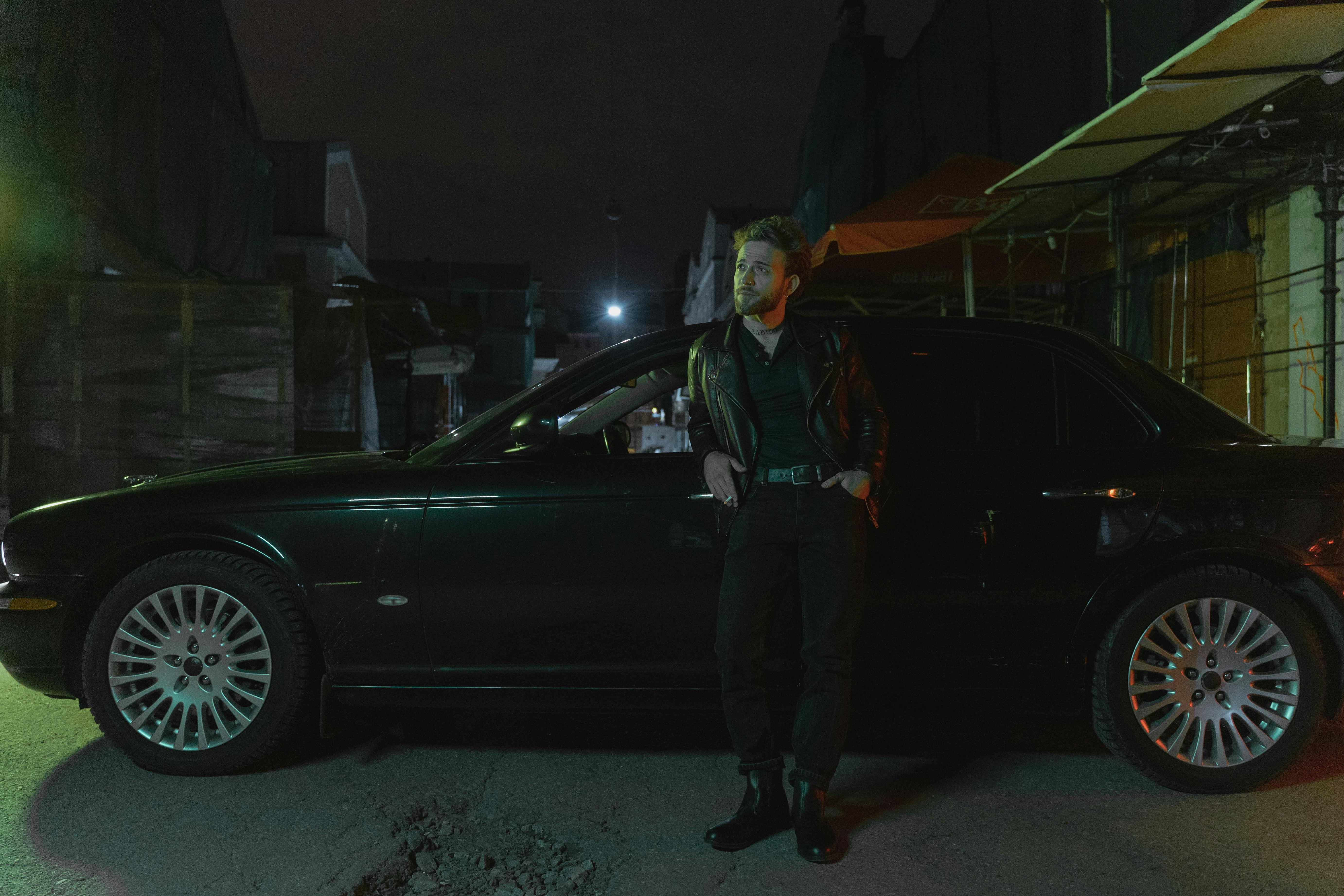 A fashionable man in a leather jacket leans on a car in an urban environment at night.