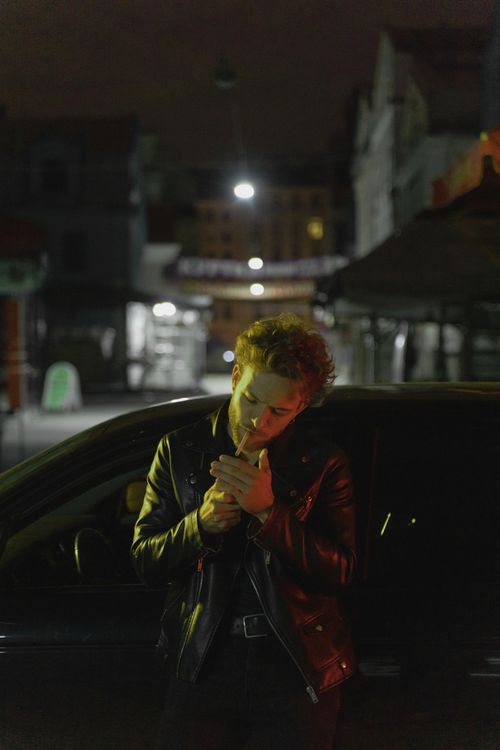 Free Man in black jacket lights a cigarette next to car in dimly lit urban area. Stock Photo