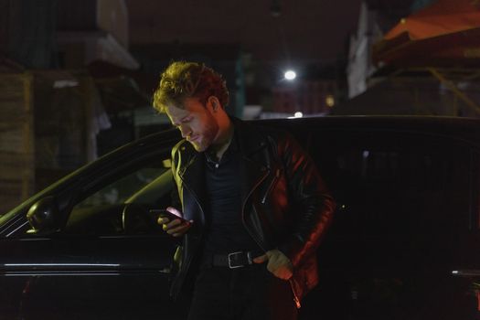 Caucasian man stands by a car at night, using his phone. Retro 80s style with a moody atmosphere.