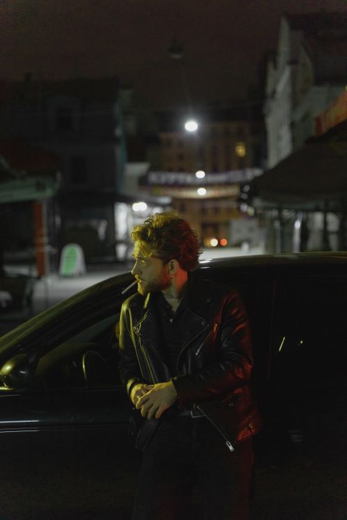 Free A man in a black leather jacket leans on a car in a city at night, smoking a cigarette. Stock Photo