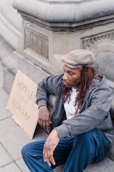 A man with braided hair sits on a sidewalk with a sign, highlighting social issues.