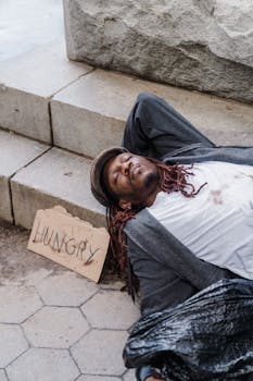 Homeless man sleeping on stairs with a cardboard sign reading 'Hungry', highlighting social issues.