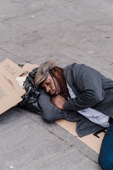 A homeless man sleeping on the street with cardboard signs and belongings.