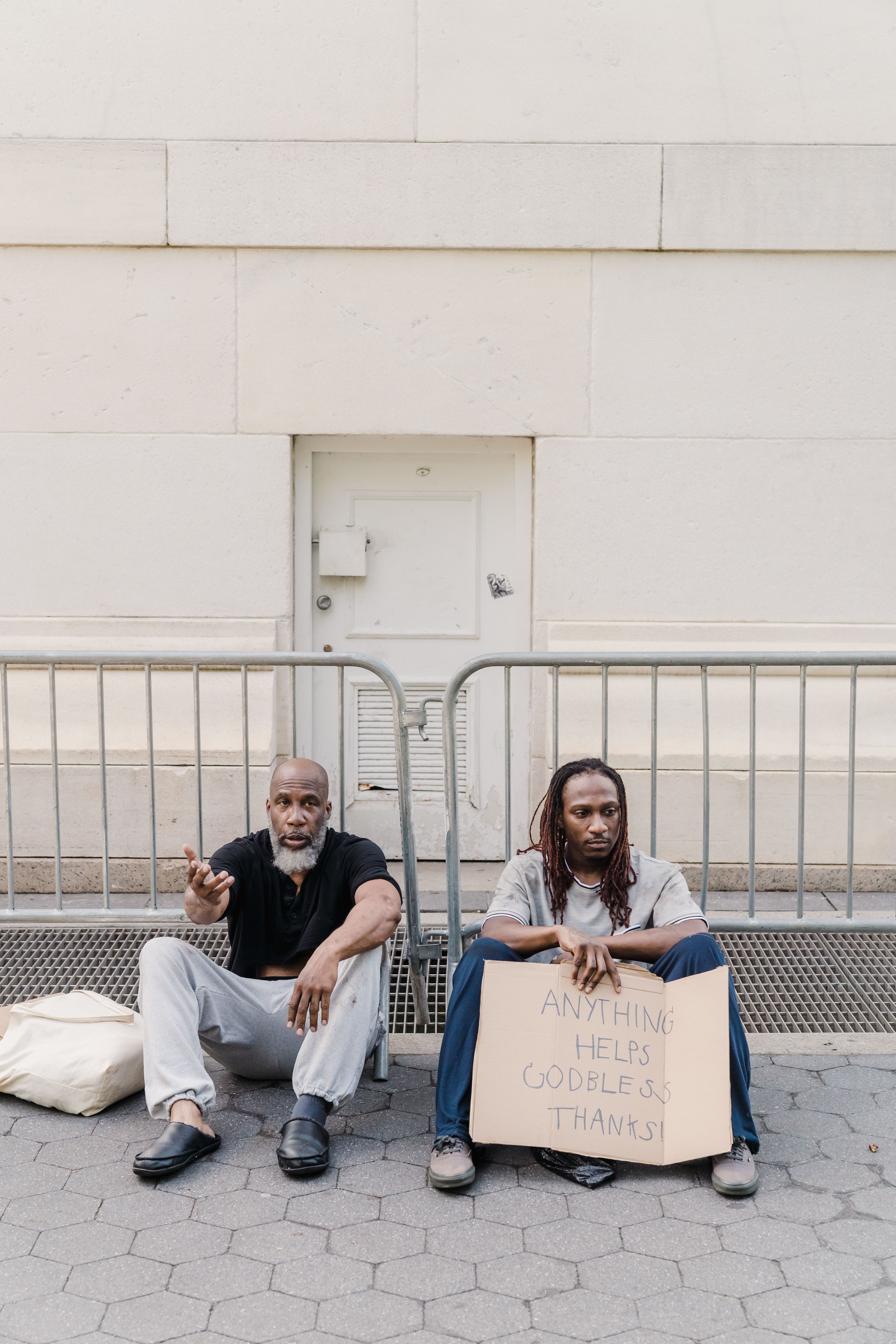 Two homeless men sitting on city sidewalk holding a sign seeking help.
