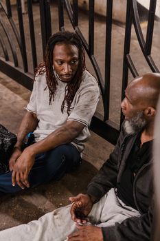 Two men engage in conversation while sitting on a sidewalk outdoors.