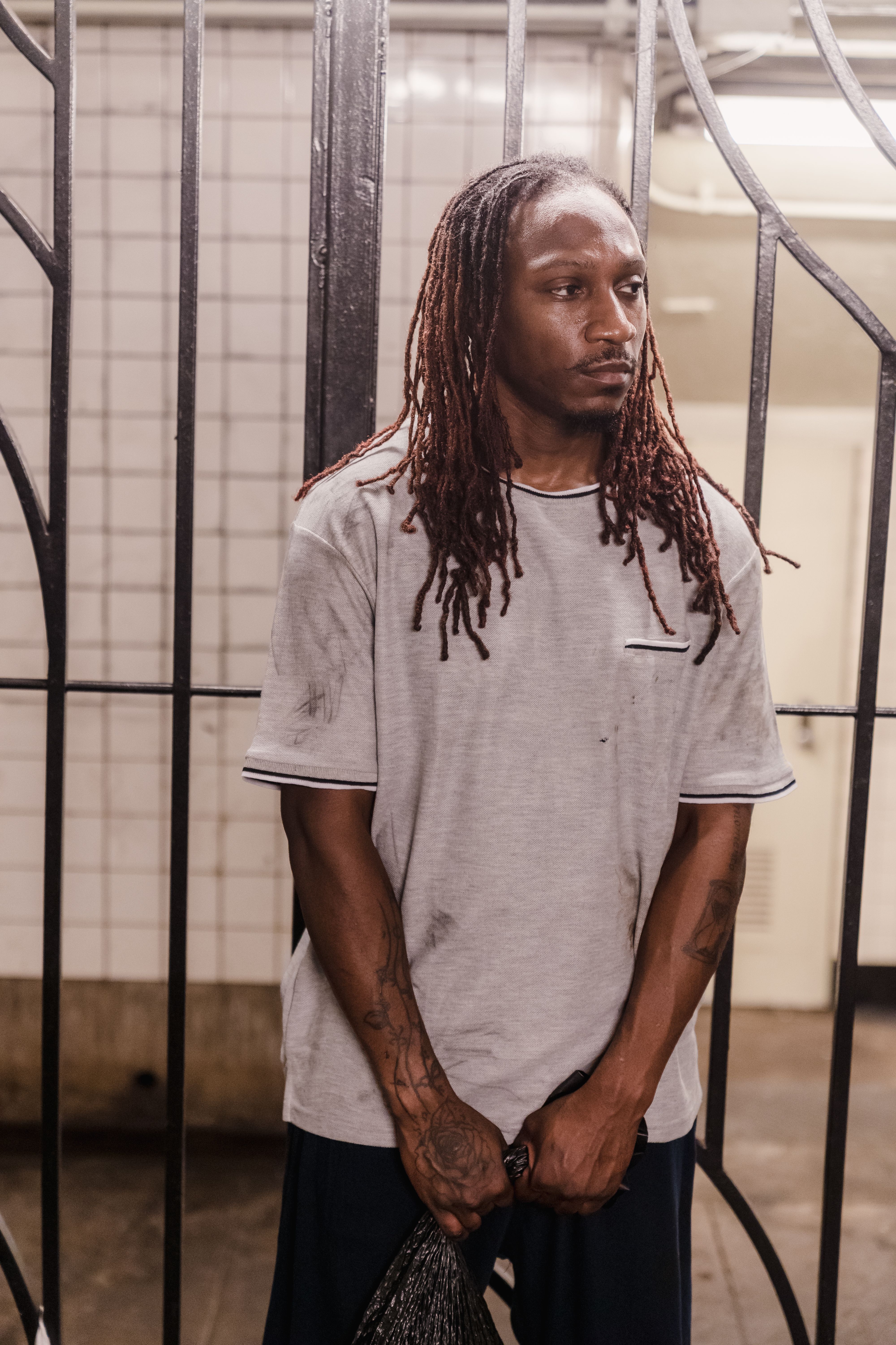 A contemplative man with dreadlocks stands indoors against iron bars, highlighting urban struggle.