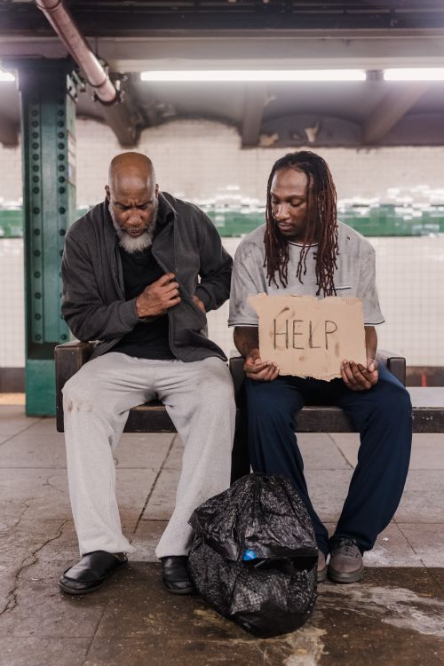 Free Two men sitting on a bench in a subway station, one holding a cardboard sign with 'HELP' written on it, highlighting homelessness. Stock Photo