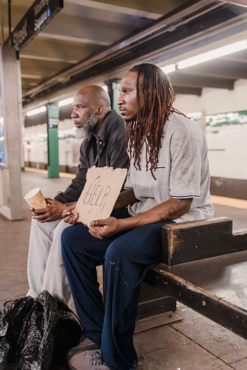Free Two men sit on a subway platform, looking pensive, holding a help sign. Stock Photo