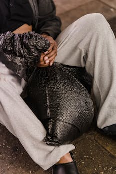 Homeless man sitting outdoors holding a black bag, highlighting urban poverty.