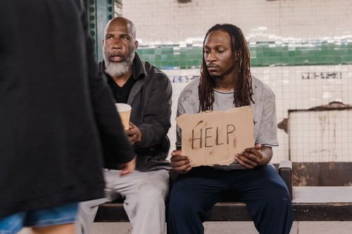 Free Two men sitting on a bench holding a help sign, symbolizing social issues and homelessness. Stock Photo
