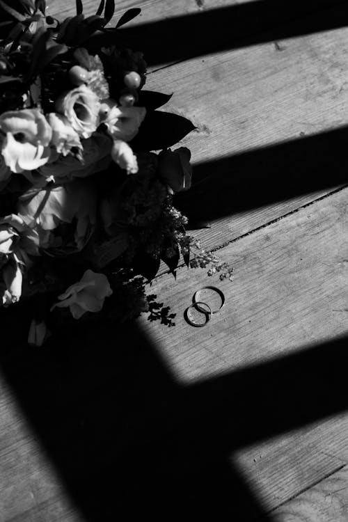 Free Grayscale Photo of Flowers Beside Wedding Rings on Wooden Surface Stock Photo