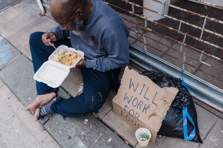 A Homeless Man Eating On A Sidewalk