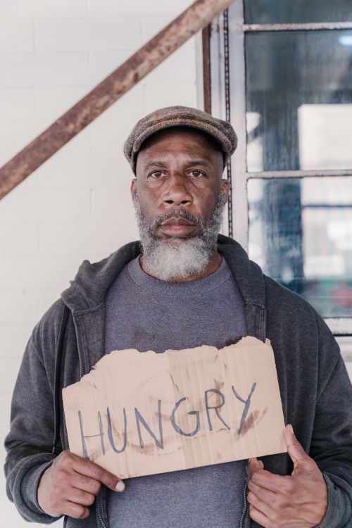 Free Older man with a beard holding a cardboard 'Hungry' sign indoors, prompting social awareness. Stock Photo