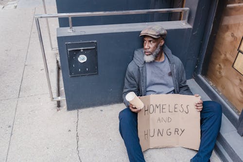 Free  Homeless Man Sitting on the Street Holding a Cup and Sign Board Stock Photo
