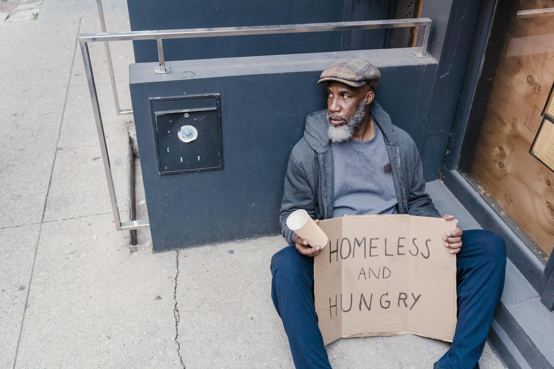 Homeless Man Sitting on the Street Holding a Cup and Sign Board