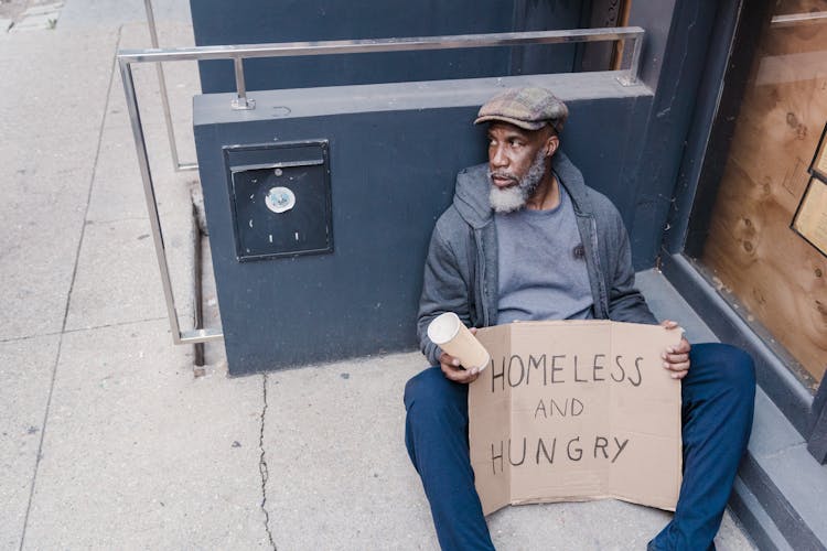  Homeless Man Sitting On The Street Holding A Cup And Sign Board