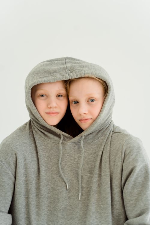 Free Two twin girls playfully share a gray hoodie against a clean white background. Stock Photo