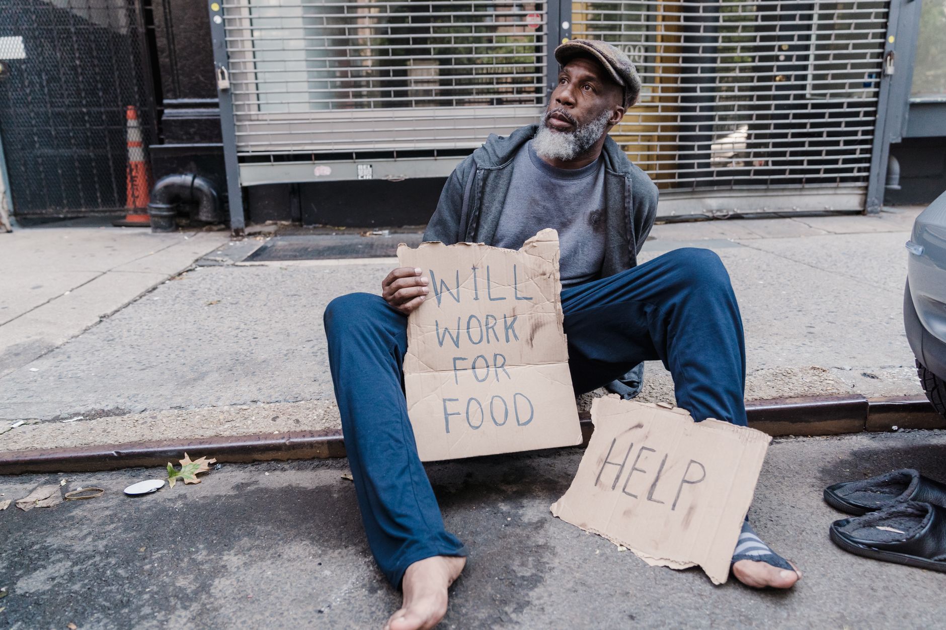 Homeless man sitting on sidewalk holding signs for help and work on a city street.