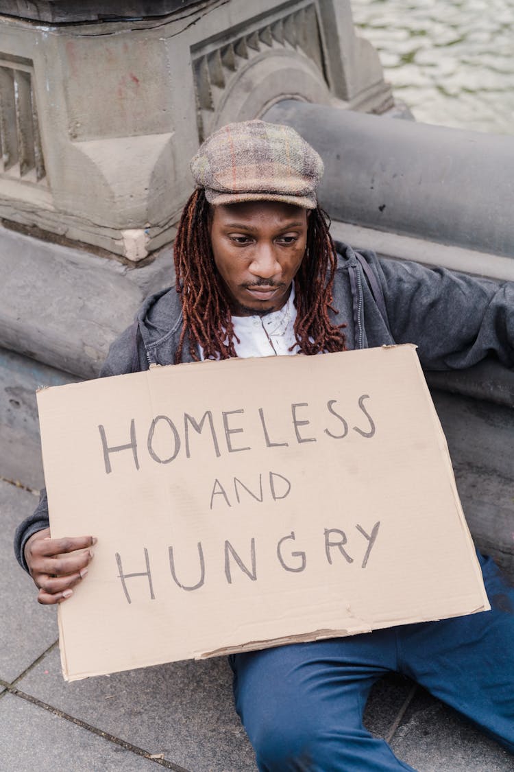 A Man Leaning On Concrete Post Sitting Holding A Cardboard Sign