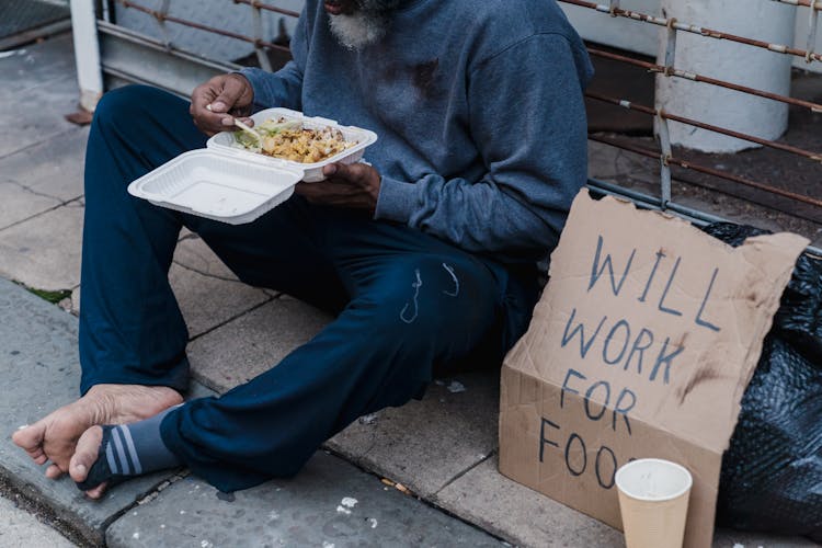 A Bearded Man Sitting On The Floor Eating Food In A Disposable Food Container