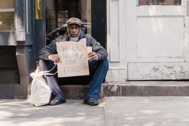 A Mature Man Sitting On The Doorway Holding A Cardboard A Sign