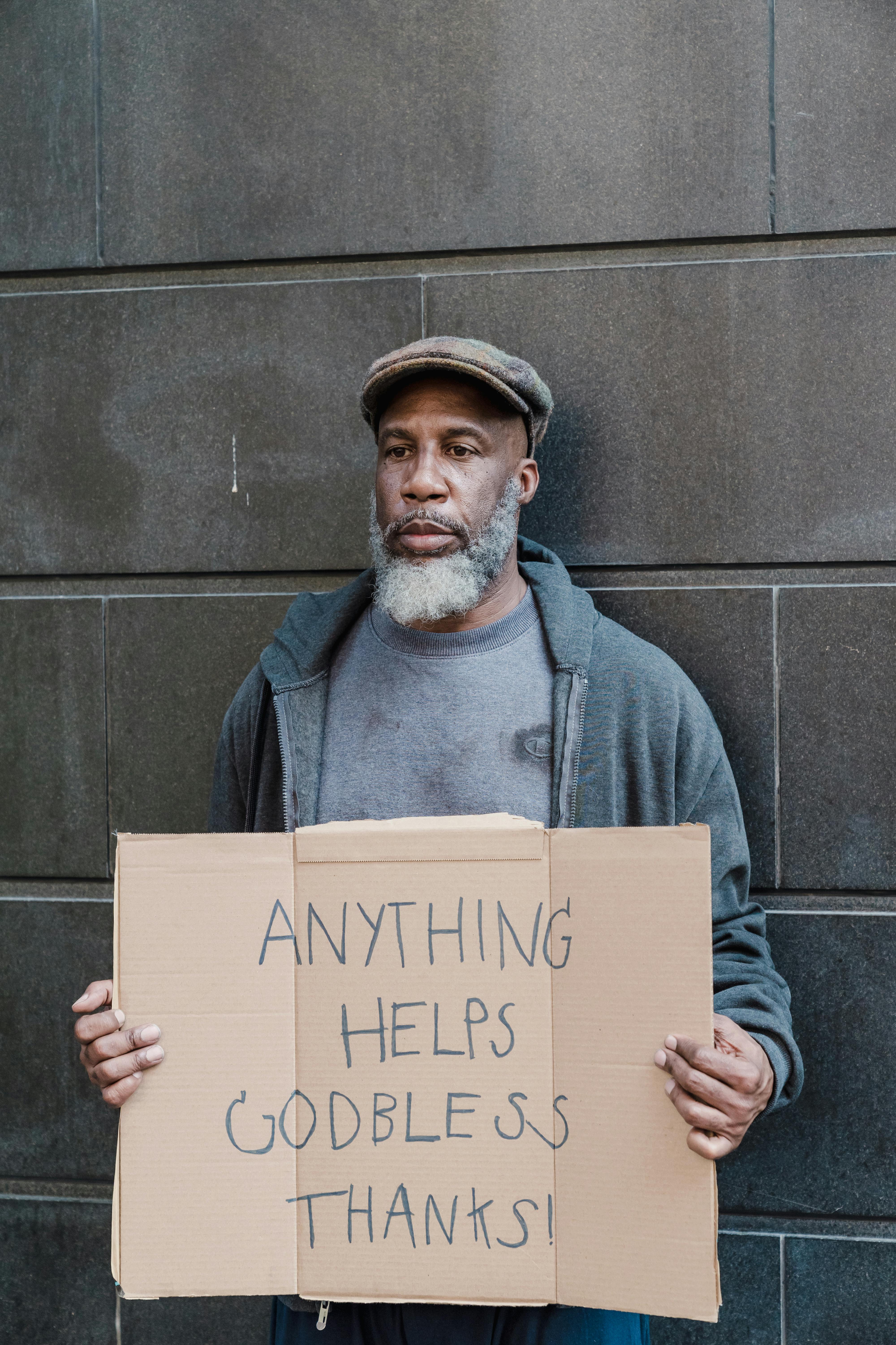Free Adult man with a beard holding a cardboard sign asking for help against a city wall. Stock Photo