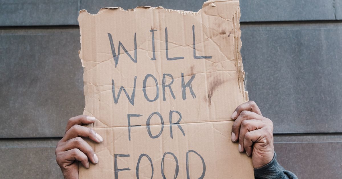 Photo by Timur Weber An adult holds a cardboard sign reading "WILL WORK FOR FOOD" in front of a gray wall, symbolizing economic hardship.