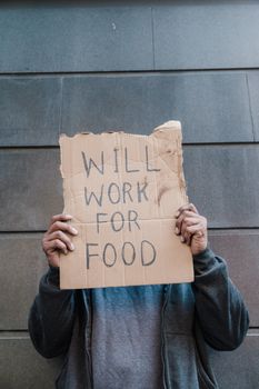 Photo by Timur Weber An adult holds a cardboard sign reading "WILL WORK FOR FOOD" in front of a gray wall, symbolizing economic hardship.