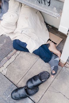 A person sleeps on the sidewalk under a blanket, highlighting homelessness and urban struggles.