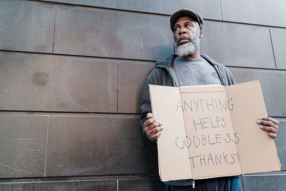 Elderly man with beard holding a handwritten sign seeking help and blessings against a stone wall.