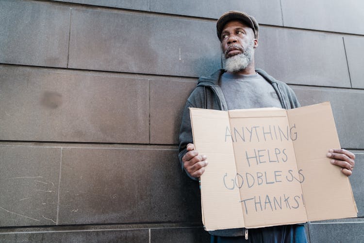 An Elderly Man Standing Beside A Wall Holding A Cardboard Sign